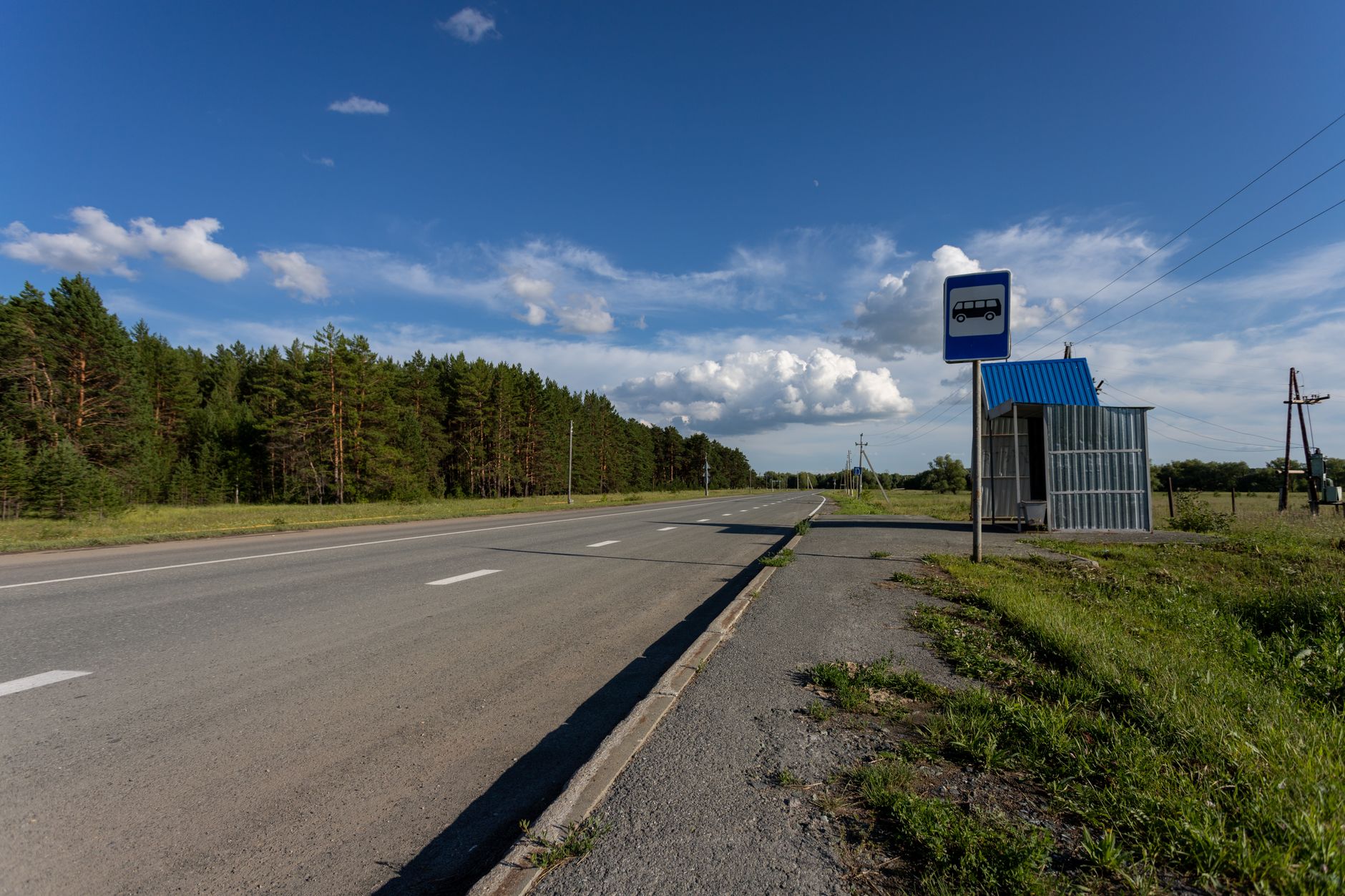 Empty rural road with a bus stop in Sinitsyna, Russia, surrounded by green trees and blue skies.