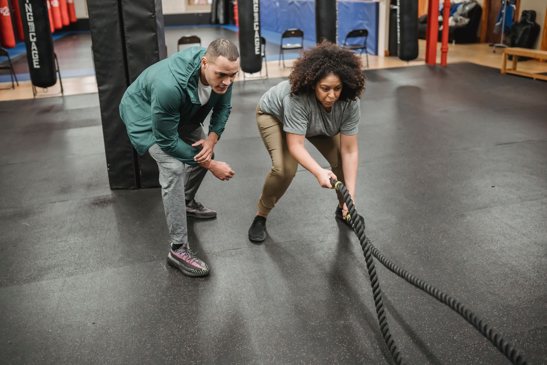 Full body focused African American female in activewear performing battle ropes exercise near fit professional male trainer in sports center