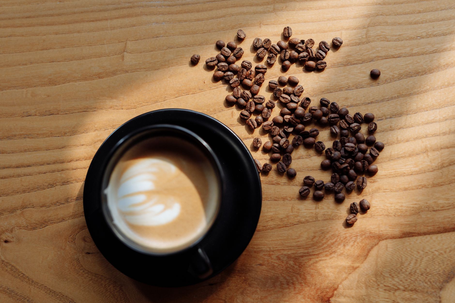 Tasse noire de café au lait avec motif de mousse, vue de dessus, et grains de café torréfiés éparpillés sur une table en bois clair.